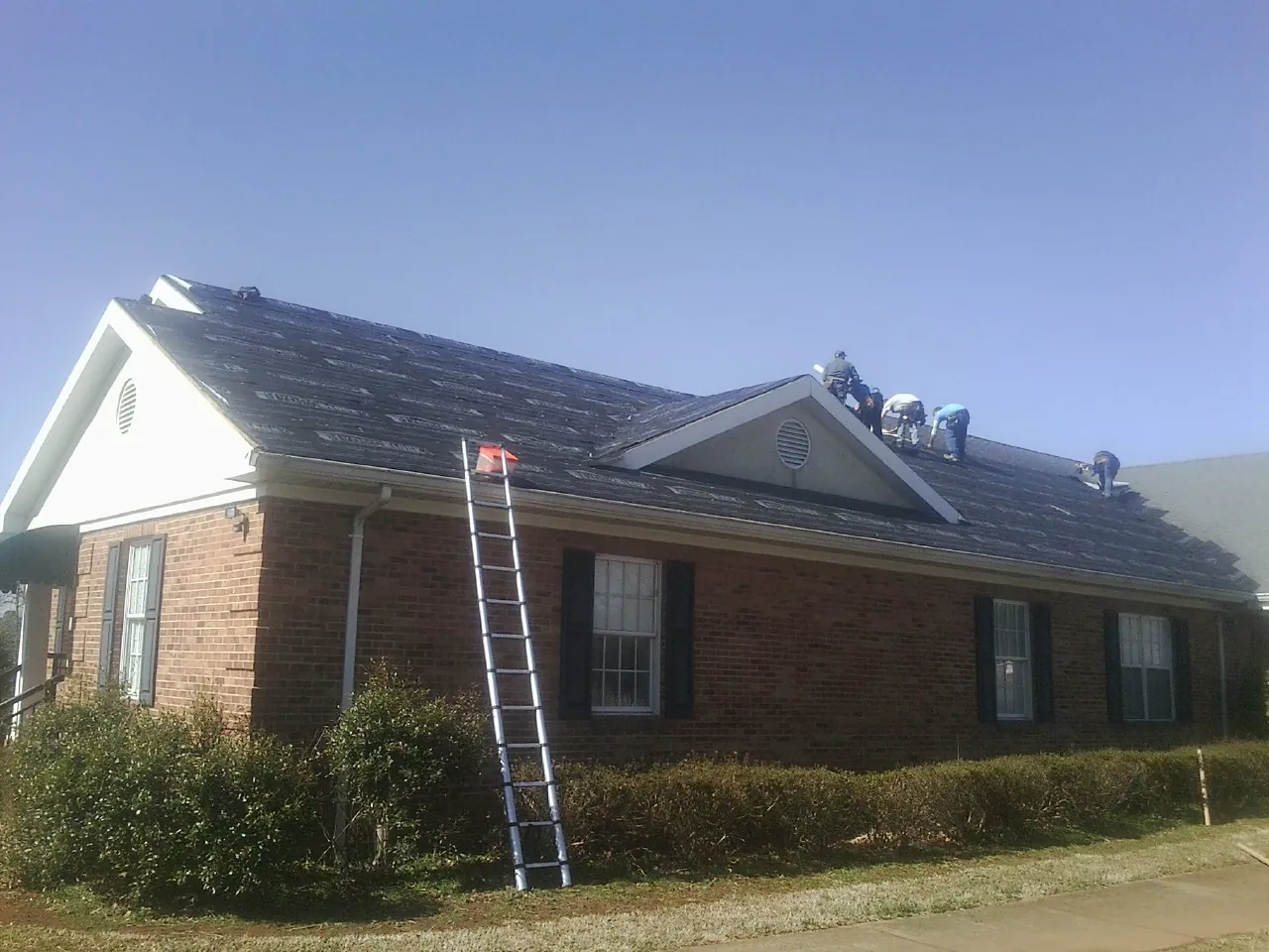 Crew tearing shingles off a brick ranch roof with a ladder set — Metal Roof Repair work by our crew in Cecilia