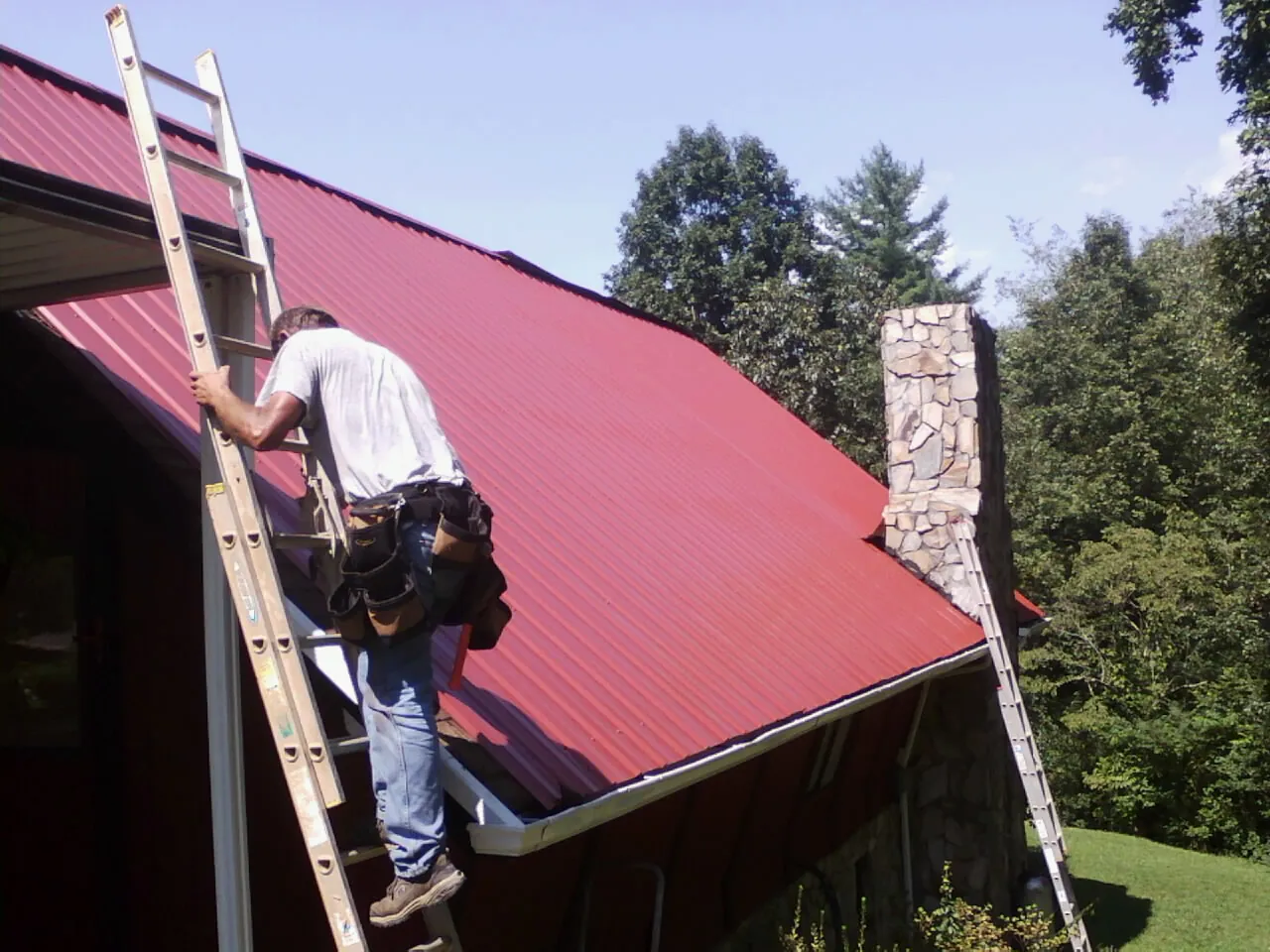 Roofer on a ladder beside a red metal roof and stone chimney — Soffit & Fascia Repair by Roofing Brothers in Cecilia